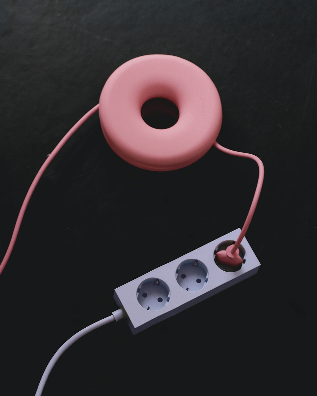 Cable Bagel in Bubble Gum su superficie scura con cavo rosa avvolto e ciabatta bianca a tre prese, vista dall'alto