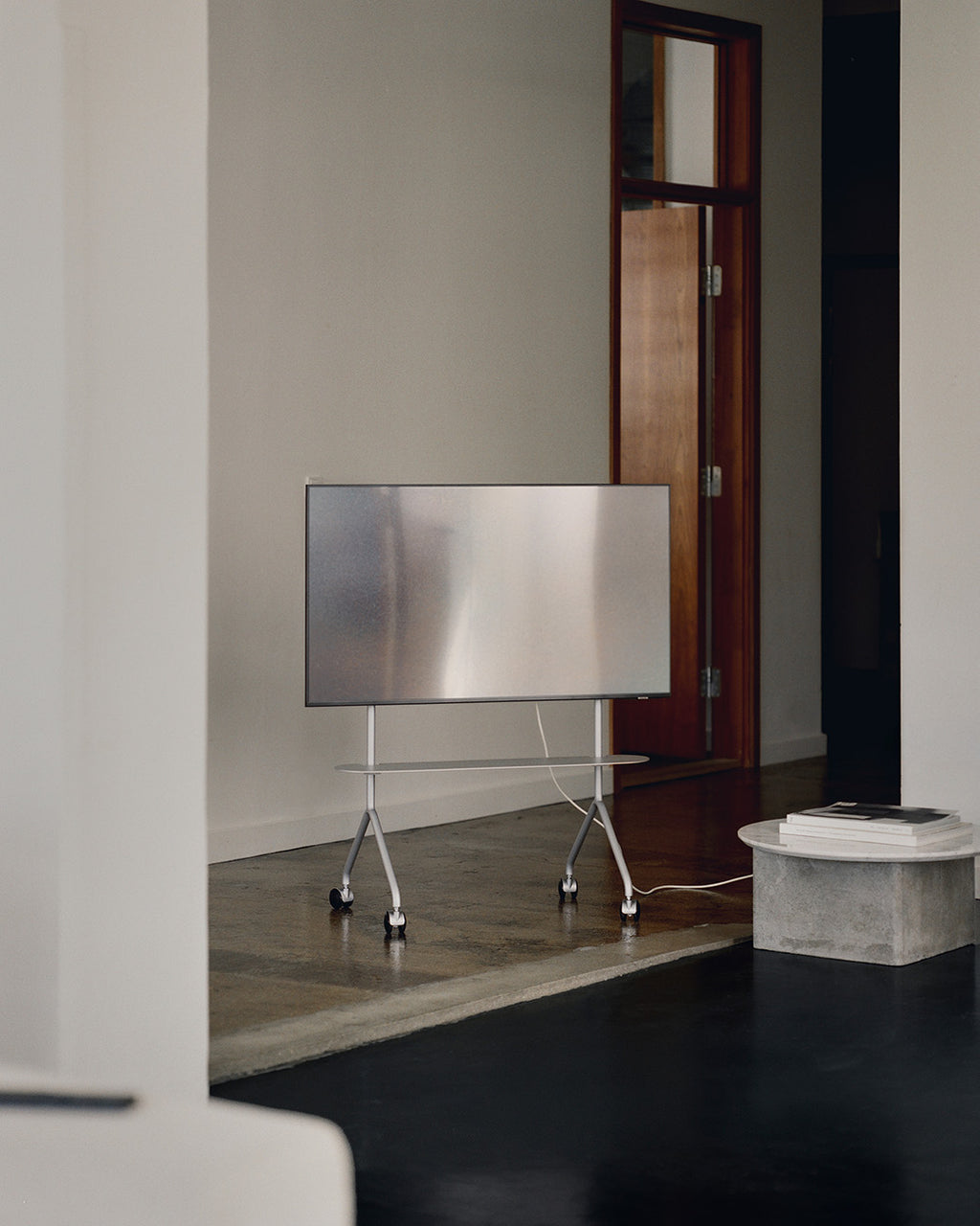 Moon Light in Cloudy Silver con TV montata e ruote, in ambiente minimal con pavimento in cemento e porta in legno